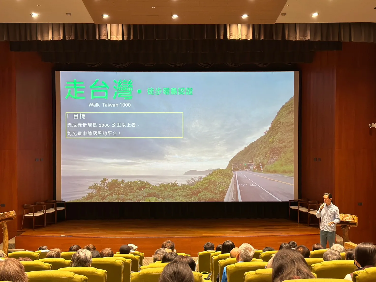 清溪文薈館的大銀幕上是「走台灣・徒步環島認證 Walk Taiwan 1000」簡報，寫著完成徒步環島 1000 公里以上者可免費申請認證，講者站在舞台右側，觀眾背對鏡頭